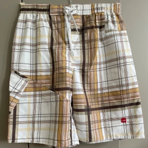 Rutiga badshorts i beige och vit - Snygga badshorts med rutigt mönster i vitt, brunt och gult. De har elastisk midja med snörning och en praktisk ficka med lock på sidan. Perfekta för stranden eller poolen, med avslappnad passform och lätt material som torkar snabbt.