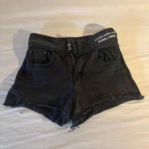 Svarta jeansshorts från SYSAN KOREA - Snygga svarta jeansshorts med mid midja och råa, fransiga kanter. Shortsen har klassiska fem fickor, dubbla knappar och vit broderad text i midjan. Perfekta för sommardagar när du vill ha en cool och avslappnad look.