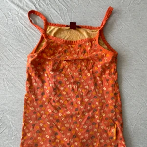 Blommig orange linnetopp - Söt orange linnetopp med små blommor i vitt, gult och mörkgrönt. Tunt och mjukt material med smala axelband, perfekt för varma dagar. Blommigt mönster ger en retrovibe och toppen är lätt att matcha med jeans eller shorts.