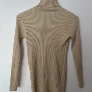 Beige ribbad polotröja - Snygg beige polotröja med ribbat material och lång ärm. Tröjan har en tight passform och hög krage som ger en clean och stilren look. Perfekt att styla med jeans eller kjol för en trendig vibe.