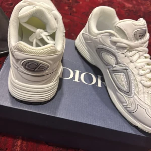 Dior B30 vit - Snygga vita Dior-sneakers med coola detaljer och CD-logga på hälen. Skorna har en chunky sula, mesh och skinnpaneler samt snörning framtill. Perfekta för dig som vill sticka ut med exklusiva och trendiga sneakers. Dior b30