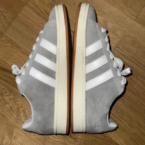 Adidas Campus 00s grå/vita sneakers - Adidas Campus 00s sneakers i ljusgrå mocka med klassiska vita ränder och vit sula. Skorna har rund tå, platt sula och breda vita skosnören. Perfekt för dig som gillar retrostil och streetwear. Snygg och tidlös modell med diskret branding på sidan. Storlek 44, passar 43. 