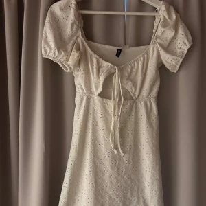 Vit kort klänning från H&M Divided XS - Supersöt vit kort klänning från H&M Divided i storlek XS. Klänningen har broderade detaljer, puffärmar och knytning framtill. Perfekt för sommarens alla tillfällen med sin luftiga bomullskänsla och romantiska look.