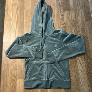 Grön velourhoodie från Juicy Couture - Mjuk och lyxig grön hoodie i velour från Juicy Couture. Tröjan har huva med snörning, dragkedja framtill och två fickor. Ikonisk JC-logga broderad på bröstet. Perfekt för en chill och trendig look.