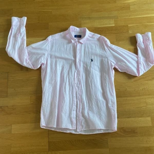 Ljusrosa linneskjorta från Polo Ralph Lauren - Klassisk ljusrosa skjorta från Polo Ralph Lauren med broderad logga på bröstet. Skjortan har lång ärm, knappar hela vägen och en tidlös passform. Perfekt för dig som gillar stilrena och fräscha plagg. Tillverkad i mjuk bomull. Perfekt för en sommar kväll Helt utan några defekter, FINNS SAMT I ANDRA FÄRGER: Mörkblå med röd logga och Ljus blå med svart logga