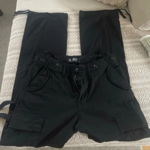 M65 Ladies Trousers | Cargo-byxor | svart | Brandit - Svarta cargobyxor från Brandit med stora fickor på sidorna, knappar i midjan och justerbara band vid benslut. Perfekta för dig som gillar streetwear och praktiska detaljer. Storlek 29 L32. Använda 1 gång