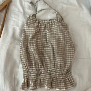 Beige och vit randig linnetopp Gina Tricot - Supersöt randig linnetopp från Gina Tricot i beige och vitt. Toppen har smala axelband som korsas i ryggen, smockad midja och volangkant nedtill. Perfekt för varma dagar och ger en chill sommarvibe. Lätt och luftig i bomullsmaterial.