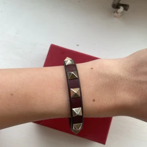 Valentino armband med nitar i läder - Snyggt armband från Valentino i mörkbrunt läder med nitar! Kommer i original ask med original dustbag! Super fint skick, lite färg-ändring + öppning av spännet! 