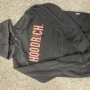 Svart hoodie från Hoodrich med tryck - Svart hoodie från Hoodrich med stort rött och vitt tryck på bröstet. Klassisk huva med snörning och känguruficka framtill. Tillverkad i mjuk bomulls- och polyestermix, perfekt för en chill och streetig look.