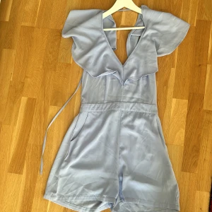 Ljusblå playsuit med volang ljusblå - Supersöt ljusblå playsuit med volangdetalj vid halsringningen och v-ringad front. Knytband i midjan och korta ben ger en avslappnad men ändå chic look. Perfekt för varma dagar när du vill känna dig både bekväm och snygg.
