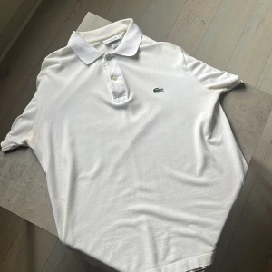 Vit pikétröja från Lacoste - Klassisk vit pikétröja från Lacoste med ikoniska krokodilloggan broderad på bröstet. Tröjan har krage, två knappar och är tillverkad i bomull. Perfekt för en clean och sportig look.