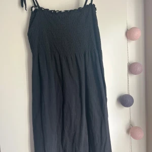 Svart sommarklänning med smock - Svart klänning med smock upptill och tunna axelband. Lätt och luftig modell som passar perfekt varma dagar. Axelbandslös känsla med enkel, rak passform och mjukt tyg. Stilren och enkel att matcha med accessoarer.