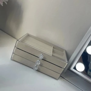 Beige smyckesförvaring med kristallknoppar - Snygg smyckesförvaring i beige sammet med tre lådor och genomskinligt lock. Perfekt för att organisera ringar, armband, halsband och örhängen. Lådorna har glittriga kristallknoppar som ger en lyxig känsla och insidan har olika fack för smycken.