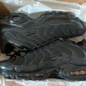 Nike Tn - Nike tn i helsvart med mesh och syntetpaneler, ikoniska vågformade detaljer och synlig Air Max-dämpning i sulan. Skorna har snörning och robust yttersula för extra grepp. Perfekta för dig som gillar en clean och sportig look.
