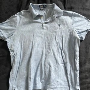 Ljusblå pikétröja från Ralph Lauren - Snygg ljusblå pikétröja från Polo Ralph Lauren i klassisk passform. Tröjan har korta ärmar, krage med två knappar och den ikoniska broderade loggan på bröstet. Perfekt för en clean och stilren look. Bara skriva vid funderingar!
