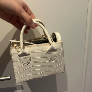 Vit croco axelväska med handtag - Säljer en liten vit axelväska med croco-mönster och både handtag och axelrem. Man kan ta bort axelremen när man inte vill ha den och sätta tillbaka den lika lätt. 