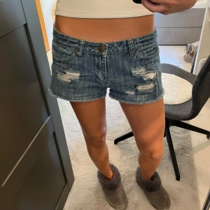 Blå slitna jeansshorts Love H81 - Snygga blå jeansshorts från Love H81 med låg midja, slitna detaljer och råa kanter. Shortsen har coola kedjedetaljer i de trasiga partierna framtill och klassisk femficksmodell. Perfekta för sommaren och ger en avslappnad vibe.