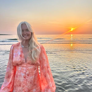 Blommig omlottklänning i rosa toner - Superfin blommig klänning i ljusrosa och orange toner med v-ringning, långa puffärmar och knappar framtill. Snygg omlottstil och flowy material, perfekt för sommarkvällar vid stranden.