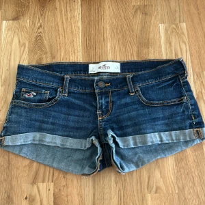 Mörkblå lågmidjade jeansshorts från Hollister - Snygga mörkblå jeansshorts från Hollister med klassisk femficksdesign, uppvikta benslut och kontrastsömmar. Shortsen har låg midja och coola detaljer på bakfickorna. Perfekta för varma sommardagar och enkel att matcha med allt.