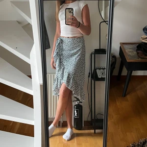 Grön blommig omlottkjol med volang - Supersöt grön omlottkjol med vitt blommönster och volangkant. Kjolen är vadlång och har knyt i sidan för att kunna reglera storleken något. Stunn och sval, perfekt till sommaren🌸