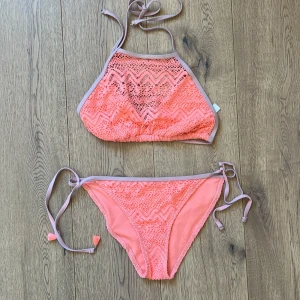 Korallrosa bikini med spets och knyt - Superfin bikini i korallrosa med spetsdetaljer och zigzag-mönster. Toppen har hög hals och knyt i nacken, medan trosan har knyt i sidorna och små tofsar. (endast ena sidan) Ljuslila kantband ger en snygg kontrast. Perfekt för stranden eller poolen!