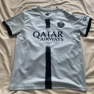 PSG fotbollströja - Snygg PSG fotbollströja i ljusblått med mörkblå detaljer och tryck. Framsidan har Qatar Airways-logga och PSG-emblem, baksidan har Messi 30-tryck. Kortärmad modell med svarta muddar och vertikal rand på framsidan. Perfekt för dig som älskar fotboll och Messi.