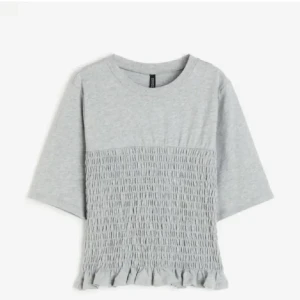 H&M smockad topp - Superfin ljusgrå topp med smockad nederdel och volangkant. T-shirten har rund halsringning och lite vidare, korta ärmar. Perfekt för dig som gillar en chill men ändå söt stil.. storlek xxs men passar mig som brukar ha xs-s