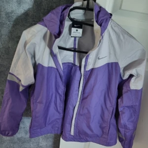 Lila och grå Nike vindjacka S - Nike vindjacka i lila och ljusgrått med huva och dragkedja framtill. Jackan har meshpaneler på sidorna för extra ventilation och reflekterande detaljer på ärmarna. Perfekt för blåsiga dagar och sportiga outfits.
