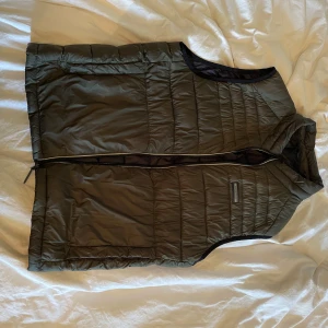 Svart quiltad dunväst med dragkedja - Säljer en mörkgrun dunväst från Jack n Jones. Västen har en stilren look med diskreta sömmar och är perfekt att slänga över en hoodie eller tröja. Materialet är lätt och smidigt, vilket gör den enkel att bära under kyliga dagar. Storleken är L men sitter mer som M eller S 