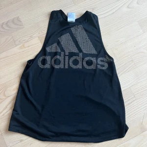 Svart linnetopp från Adidas - Snygg svart linnetopp från Adidas med det ikoniska logotyptrycket i silver. Perfekt för träning eller en avslappnad dag. Toppen har en lös passform och är ärmlös för maximal rörelsefrihet.