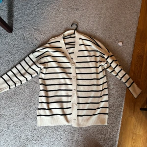 Randig kofta i beige och svart - Säljer en stilren randig kofta i beige med svarta ränder. Koftan har långa ärmar och knappar framtill. Perfekt för en avslappnad look.