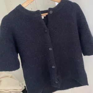 Mörkblå stickad kofta - Säljer en mysig mörkblå stickad kofta med korta ärmar och knappar framtill. Perfekt för kyliga dagar och ger en stilren look. Passar bra till både jeans och kjol.