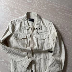 Beige jacka från Emporio Armani strl: 46 - Säljer en stilren beige jacka från Emporio Armani. Jackan har dragkedja och flera praktiska fickor framtill. Perfekt för en avslappnad och trendig look. Passar bra till både sommar och  vår. 
