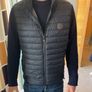 Mörkblå väst från Marc O'Polo - Snygg svart quiltad väst från Marc O'Polo med dragkedja framtill. Västen är tillverkad med termisk isolering från Dupont™ Sorona® som är ett alternativ till dun och fjädrar. Perfekt för att hålla dig varm utan att vara tung.