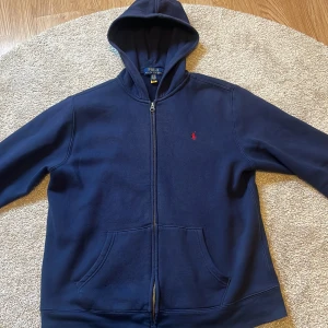 Mörkblå hoodie från Ralph Lauren - Säljer en snygg mörkblå hoodie från Ralph Lauren med dragkedja och huva. Den har en liten röd logga på bröstet och praktiska fickor framtill. Perfekt för en avslappnad stil. Den är i jättebra skick och pris går att diskutera med rimliga gränser🙌 det är storlek XL i ungdom men skulle säga ungefär S i herrstorlek, hör av dig om passningsform om du vill