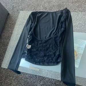 Svart långärmad topp med spetsdetaljer - Snygg svart långärmad topp med spetsdetaljer framtill. Perfekt för en elegant look med en unik touch. Toppen har en rund halsringning och är tillverkad i ett mjukt material.