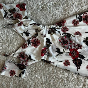 Vit blommig långklänning - Säljer en vacker vit långklänning med blommigt mönster i rött, rosa och grönt. Klänningen har en omlott-design och korta ärmar med en elegant v-ringning. Perfekt för sommarens festligheter!