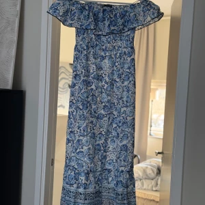 Blåvit blommig långklänning - Charmig blåvit långklänning med blommigt mönster och volangdetalj vid axlarna. Perfekt för somriga dagar med sin luftiga design och offshoulder-stil.