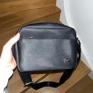 Svart axelväska från Lacoste - Snygg svart axelväska från Lacoste med en stilren design. Väskan har en justerbar axelrem och en praktisk dragkedja framtill. Perfekt för att bära med sig det nödvändigaste med stil.