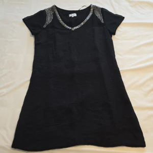 Svart klänning med pärldetaljer - Elegant svart klänning från Inwear med v-ringning och korta ärmar. Klänningen har vackra pärldetaljer vid axlarna och halsringningen, vilket ger en lyxig touch. Perfekt för en stilfull kväll.