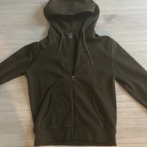 Olivgrön kofta från Ralph Lauren - Säljer en stilren olivgrön kofta från Ralph Lauren med dragkedja och justerbar huva. Tröjan har en broderad logga på bröstet och praktiska fickor framtill. Perfekt för en avslappnad look.
