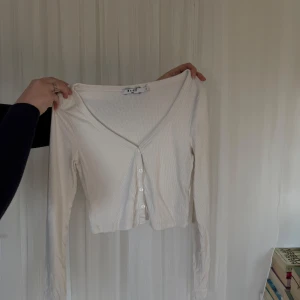 Vit ribbad kofta från NAKD - Säljer en stilren vit ribbad kofta från Nakd. Den har en v-ringad design med knappar framtill och långa ärmar. Perfekt för lager-på-lager-stil eller som ett lättare plagg under kyligare dagar.