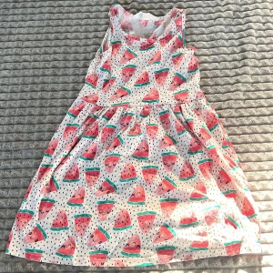 Vit klänning med vattenmelonmönster - Söt vit klänning med ett lekfullt mönster av vattenmeloner. Klänningen är ärmlös och har en luftig passform, perfekt för varma sommardagar. Den har en rund halsringning och är gjord i ett lätt och bekvämt material.