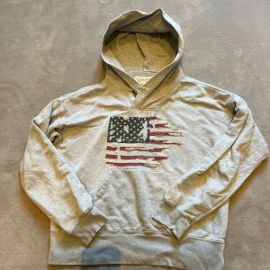 Grå hoodie från Ralph Lauren - Säljer en grå hoodie från Ralph Lauren med ett coolt amerikanskt flaggmotiv på framsidan. Hoodien har en knappdetalj vid halsen och är tillverkad i mjuk bomull. Perfekt för en avslappnad stil!