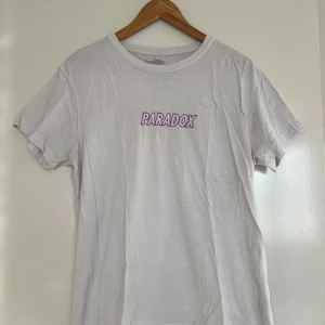 Vit t-shirt från Paradox - Säljer en vit t-shirt från Paradox med lila text på bröstet. T-shirten har en klassisk rund hals och korta ärmar. Perfekt för skate-stil.