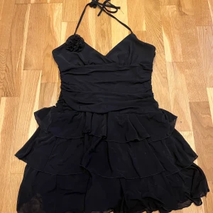 Svart klänning med volanger - Snygg svart kort klänning med volanger och band som man knycker i nacken. Klänningen har en v-ringad urringning och en dekorativ blomdetalj vid axeln. Perfekt för en elegant kväll.🖤