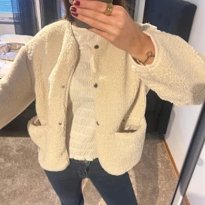Beige teddyjacka - Mysig beige teddyjacka med knappar framtill. Perfekt för kyliga dagar med sin fluffiga och varma design. Jackan har en avslappnad passform och två praktiska fickor. Passar perfekt till våren☀️💕