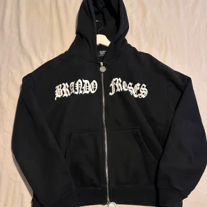 Svart hoodie från Brando Roses - Säljer en svart hoodie från Brando Roses med vit text på bröstet. Den har en dragkedja framtill och en praktisk huva. Perfekt för en avslappnad stil.