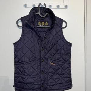 Mörkblå quiltad väst från Barbour - Säljer en stilren mörkblå quiltad väst från Barbour. Västen har en klassisk design med knappar och broderad logotyp på vänster ficka. Perfekt för en lite kallare dag man vill ha på sig nått bekvämt som värmer.