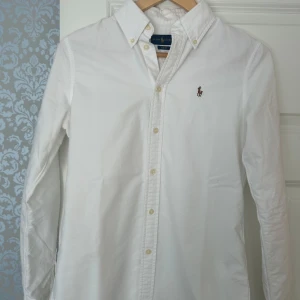 Vit skjorta från Ralph Lauren - Hjälper min lillebror sälja en stilren vit skjorta från Ralph Lauren med broderad logotyp på bröstet. Skjortan har en slim passform och är tillverkad i bomull. Perfekt för en elegant look. Skick är superbra, han säljer endast för han växt ur den. 